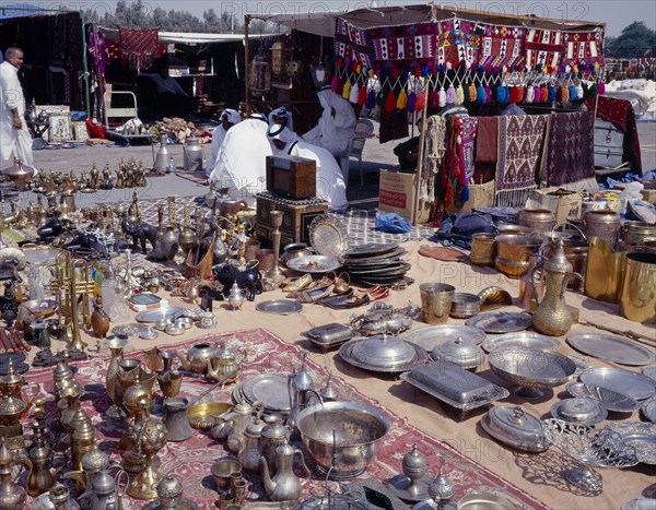 KUWAIT, Kuwait City, "Brassware, crafts and textiles for sale at the Friday Market or souq with seated men wearing Dishdasha and Gutra."