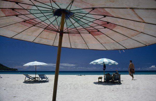 THAILAND, Phuket, Western tourists on white sand beach with sun loungers part framed by umbrella in the foreground.