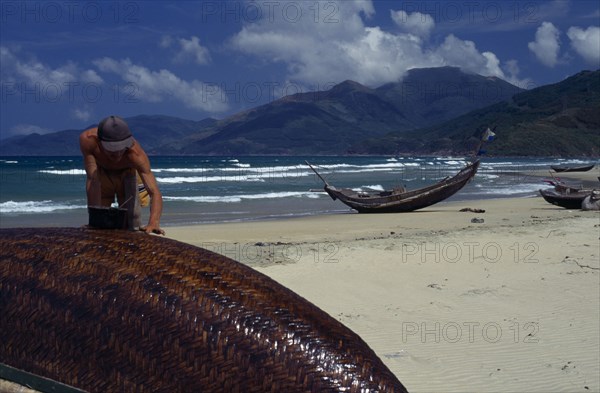 VIETNAM, Lang Co, Man sealing hull of overturned fishing boat with other boats pulled up onto sand behind.