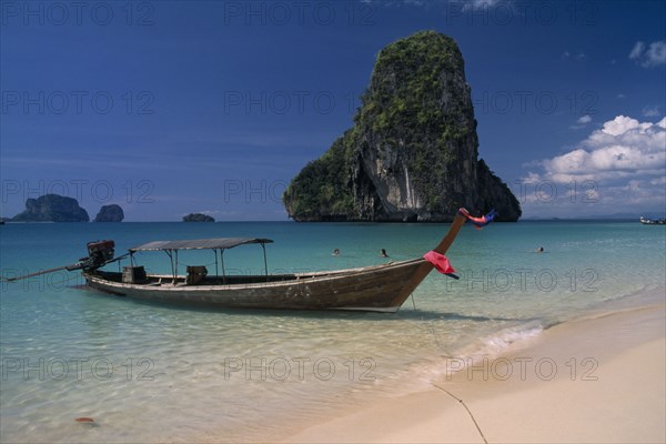 THAILAND, Krabi Province, Ao Phra Nang, Longtail boat moored in shallow water beside golden sand beach with people swimming and limestone karst rock formation behind.