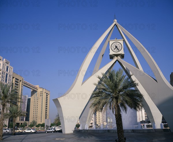 UAE, Dubai, Dubai Clock Tower.  Modern sculptural structure with pool and fountains below and palm tree in foreground.  Traffic and city buildings behind.