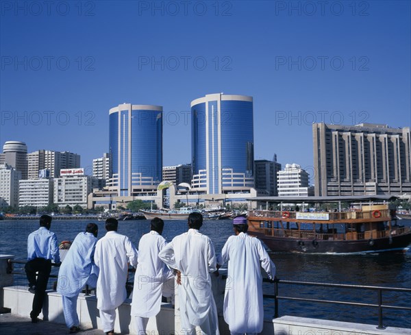 UAE, Dubai, Dubai Creek.  Men standing on waterfront looking out towards city skyline and passing dhow.