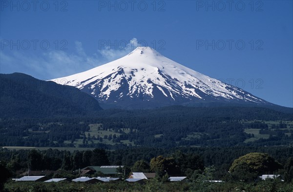 CHILE, Araucania Region, Parque Nacional Villarica , "Pucon.  Villarica Volcano 2847m.  Active volcano in the Chilean Andes, snow covered cone rising above forest covered hills with building rooftops in the foreground. "