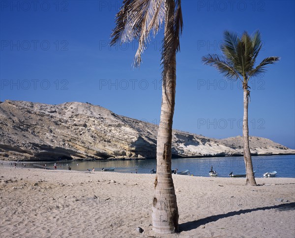 OMAN, Muscat, "Bandar Jissah beach near Al-Bustan Hotel just outside the capital city.  People at water’s edge with eroded, sloping rock formation behind and palm in foreground."