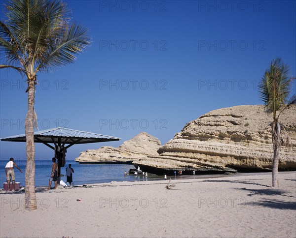 OMAN, Muscat, "Bandar Jissah beach near Al-Bustan Hotel just outside the capital city.  People at water’s edge beside eroded, rock formation with sun umbrella and palm trees."