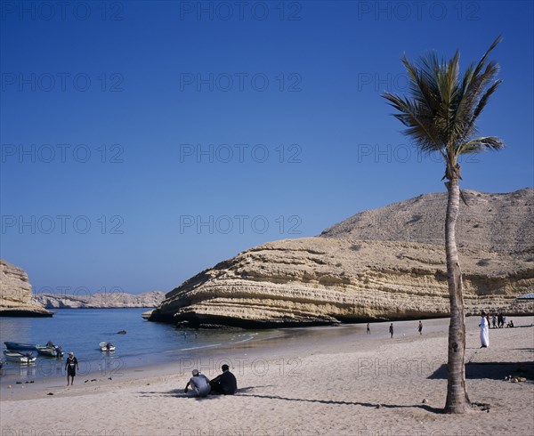 OMAN, Muscat, "Bandar Jissah beach near Al-Bustan Hotel just outside the capital city.  People at water’s edge beside eroded, rock formation with palm in foreground."
