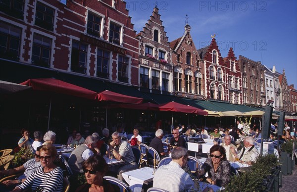 BELGIUM, West Flanders, Bruges, The Markt (Market Place) Line of cafes with busy outside tables under green awnings and red umbrellas.