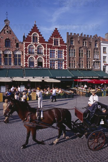 BELGIUM, West Flanders, Bruges, The Markt (Market Place).  Horse drawn carriage passing line of cafes with busy outside tables under green awnings and red umbrellas.