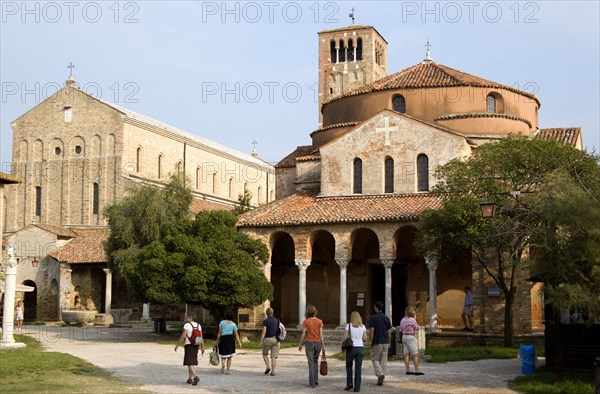 ITALY, Veneto, Venice, The uninhabited island of Torcello with the Byzantine cathedral on the left and the church of Santa Fosca on the right. Tourists walk past the front of the church