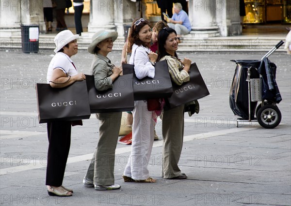 ITALY, Veneto, Venice, Asian tourists in St Marks Square posing for photographs whilst shouldering shopping bags from the nearby Gucci shop