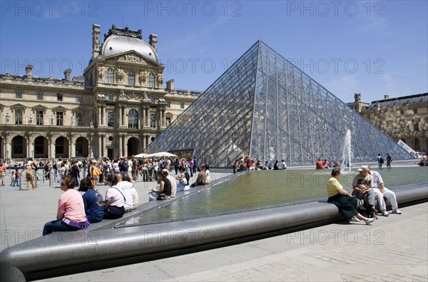 FRANCE, Ile de France, Paris, Tourists sitting beside the fountain pools in the Cour Napoleon at the Musee du Louvre with the Pyramid Entrance designed by I M Pei and the Richelieu Pavilion beyond