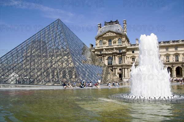 FRANCE, Ile de France, Paris, Tourists sitting beside the fountain pools in the Cour Napoleon at the Musee du Louvre with the Pyramid Entrance designed by I M Pei and the Richelieu Pavilion beyond