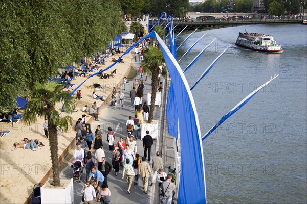 FRANCE, Ile de France, Paris, The Paris Plage city beach on the right bank of the River Seine where the road is closed and people lie in the sand and walk along the river bank. A bateaux mouches vedettes with sightseers passes under Pont D'Arcole