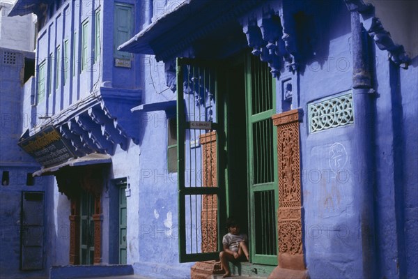 INDIA, Rajasthan, Jodhpur, Detail of blue painted house with child sitting in open doorway with green frame and decorated surround.