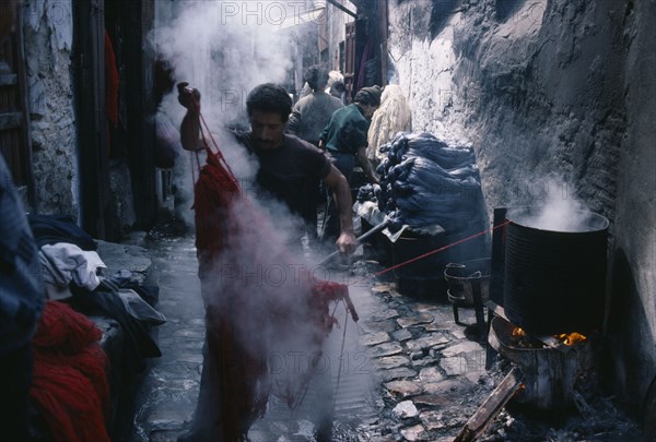 MOROCCO, Fez, Street in the wool dyeing souk with man in foreground lifting skein of steaming yarn from vat of dye.