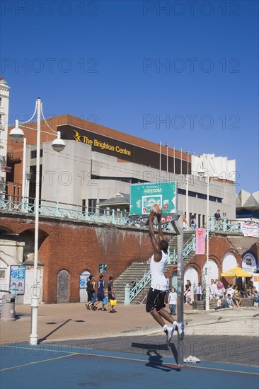 ENGLAND, East Sussex, Brighton, Young man playing basketball on the seafront court with the Brighton Centre behind.