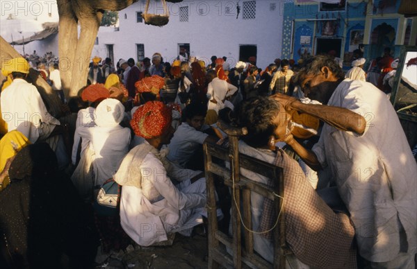 20082707 INDIA Rajasthan Pushkar Crowds of people with barber shaving customer on street in foreground.