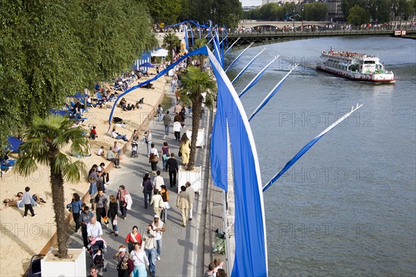 FRANCE, Ile de France, Paris, The Paris Plage urban beach. People strolling between the River Seine and other people lying on sand along the Voie Georges Pompidou usually a busy road now closed to traffic. A Pleasure boat on the river with tourists passes under the Pont D'Arcole bridge