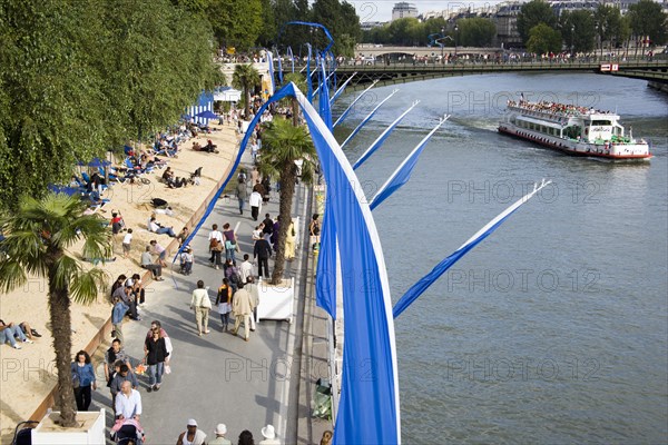 FRANCE, Ile de France, Paris, The Paris Plage urban beach. People strolling between the River Seine and other people lying on sand along the Voie Georges Pompidou usually a busy road now closed to traffic. A Pleasure boat on the river with tourists passes under the Pont D'Arcole bridge