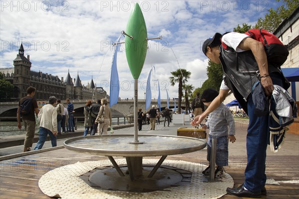 FRANCE, Ile de France, Paris, "The Paris Plage urban beach. A small boy with an adult at one of the many free drinking water fountains on the Voie Georges Pompidou, a usually busy road now closed to traffic, opposite the Ile de la Cite "
