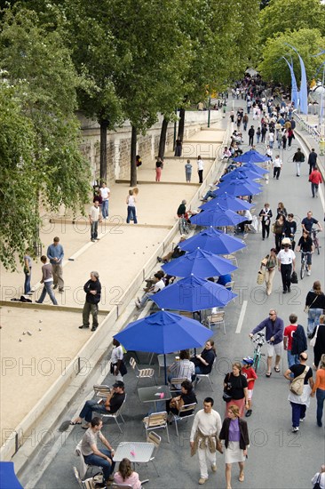 FRANCE, Ile de France, Paris, The Paris Plage urban beach. People strolling between the River Seine and people playing boules or petanque divided by a line of tables under umbrellas along the Voie Georges Pompidou usually a busy road now closed to traffic opposite the Ile de la Cite