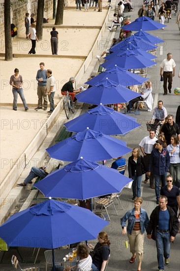 FRANCE, Ile de France, Paris, The Paris Plage urban beach. People strolling between the River Seine and people playing boules or petanque divided by a line of tables under umbrellas along the Voie Georges Pompidou usually a busy road now closed to traffic opposite the Ile de la Cite