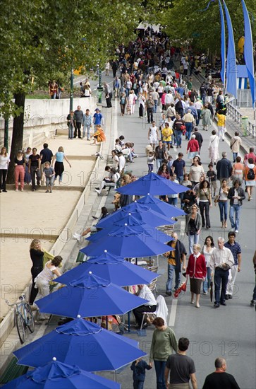 FRANCE, Ile de France, Paris, The Paris Plage urban beach. People strolling between the River Seine and people playing boules or petanque divided by a line of tables under umbrellas along the Voie Georges Pompidou usually a busy road now closed to traffic opposite the Ile de la Cite