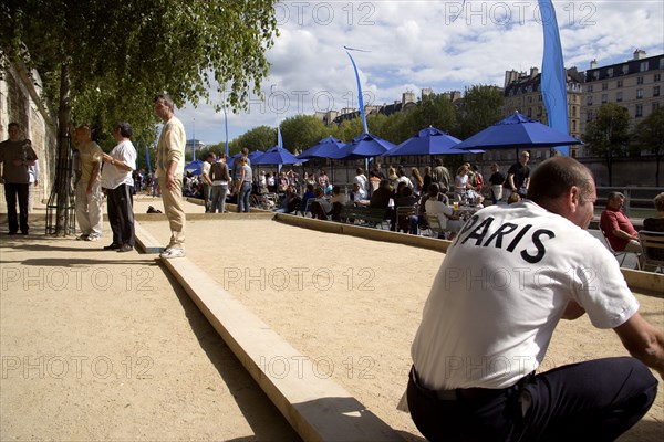 FRANCE, Ile de France, Paris, The Paris Plage urban beach. A man wearing a Paris t-shirt beside people playing boules petanque along the Voie Georges Pompidou a usually busy road closed to traffic opposite the Ile de la Cite