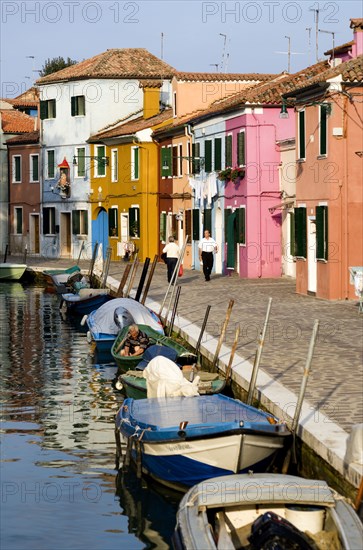 ITALY, Veneto, Venice, Colourful houses beside a canal on the lagoon island of Burano with people walking past boats moored alongside the edge of the canal