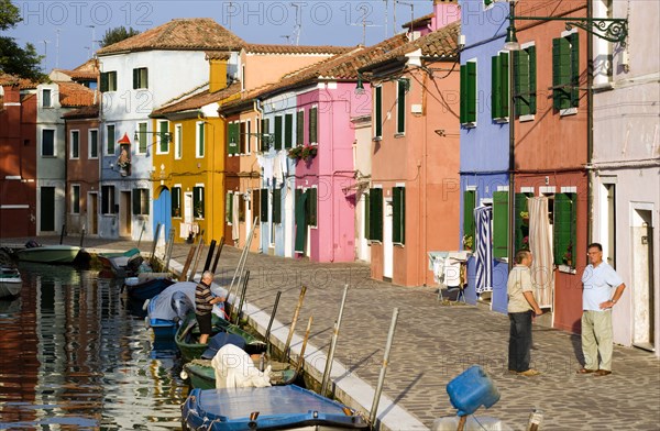 ITALY, Veneto, Venice, Colourful houses beside a canal on the lagoon island of Burano with people walking past boats moored alongside the edge of the canal