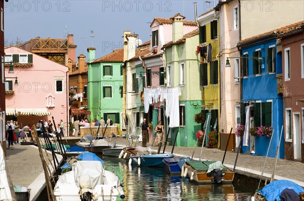 ITALY, Veneto, Venice, Colourful houses beside a canal on the lagoon island of Burano with people walking past boats moored alongside the edge of the canal. Tourists sit at tables outside a restaurant whilst others walk past shops by a bridge.