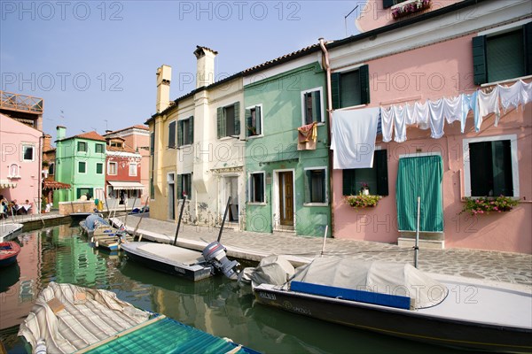 ITALY, Veneto, Venice, Colourful houses beside a canal on the lagoon island of Burano with boats moored alongside the edge of the canal. Tourists sit at tables outside a restaurant whilst others walk past shops by a bridge. Laundry hangs on a line below windows of a building
