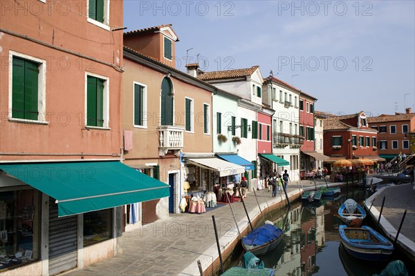 ITALY, Veneto, Venice, Colourful houses beside a canal on the lagoon island of Burano with boats moored alongside the edge of the canal. Tourists sit at tables outside a restaurant whilst others walk past shops by a bridge.