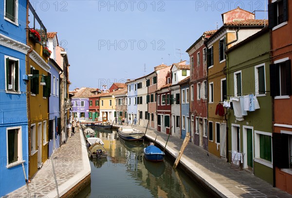 ITALY, Veneto, Venice, Colourful houses beside a canal on the lagoon island of Burano with tourists walking past boats moored alongside the edge of the canal.