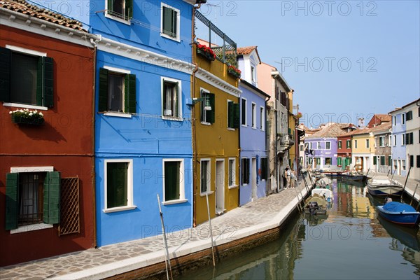 ITALY, Veneto, Venice, Colourful houses beside a canal on the lagoon island of Burano with tourists walking past boats moored alongside the edge of the canal.