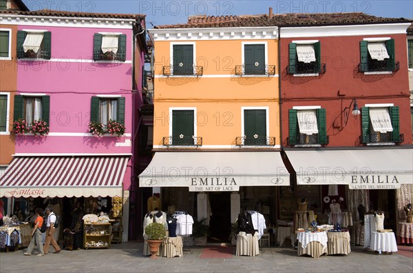 ITALY, Veneto, Venice, Brightly coloured houses above lace shops on the lagoon island of Burano the historic home of the lace making industry in the region. Tourists walk past looking at the displays on the pavement