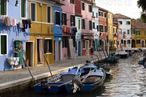 ITALY, Veneto, Venice, Colourful houses beside a canal on the lagoon island of Burano with tourists walking past boats moored alongside the edge of the canal