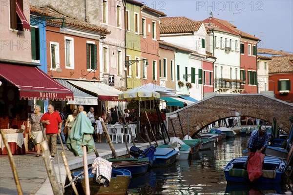 ITALY, Veneto, Venice, Colourful houses beside a canal on the lagoon island of Burano with people walking past shops and boats moored alongside the edge of the canal near to a pedestrian bridge
