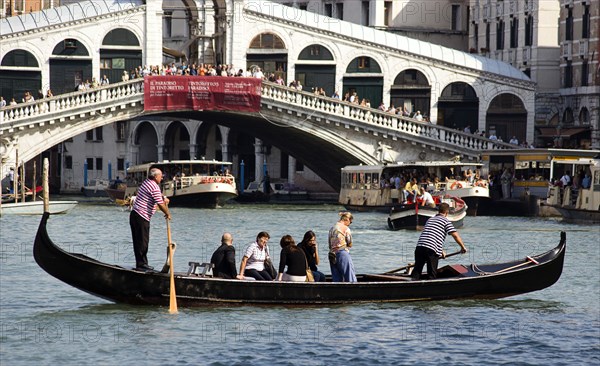 ITALY, Veneto, Venice, A Traghetto carrying local people on board crosses the Grand Canal. Tourists crowd on the Rialto Bridge spanning the canal with vaporetto passing beneath