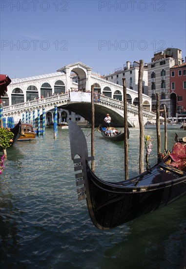 ITALY, Veneto, Venice, A gondola moored at the edge of the Grand Canal with another carrying tourists passes down river in front of The Rialto Bridge with tourist crowds