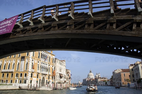 ITALY, Veneto, Venice, The Byzantine church of Santa Maria della Salute with water taxis and a motoscafo passing along the Grand Canal towards Palazzo Franchetti Cavali decked with flags. People walk across the wooden Accademia bridge spanning the canal