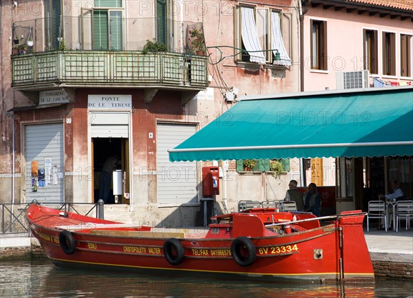 ITALY, Veneto, Venice, A traditional red lagoon barge moored alongside the Fondamenta Sebastiano Santi beside a grocery shop and coffee shop by the Ponte de la Terese bridge across the Canale di San Donato on the island of Murano
