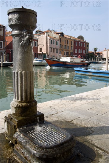 ITALY, Veneto, Venice, A free public drinking water fountain on the Canale di San Donato on the island of Murano. One of many on the island. A lagoon barge passes down the canal