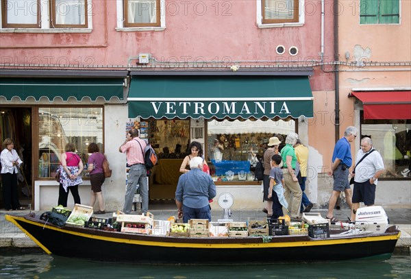 ITALY, Veneto, Venice, A fruit and vegetable vendor with his boat moored alongside the Fondamenta dei Vetrai olong the Rio dei Vetrai canal on the lagoon island of Murano. Tourists walk along the pavement looking at the shop windows