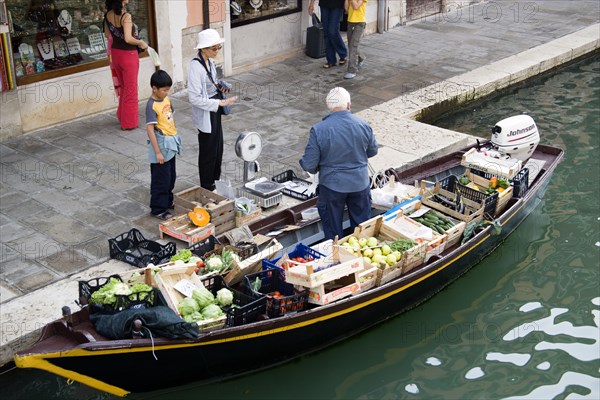 ITALY, Veneto, Venice, A fruit and vegetable vendor serving a customer from his boat moored alongside the Fondamenta dei Vetrai along the Rio dei Vetrai canal on the lagoon island of Murano