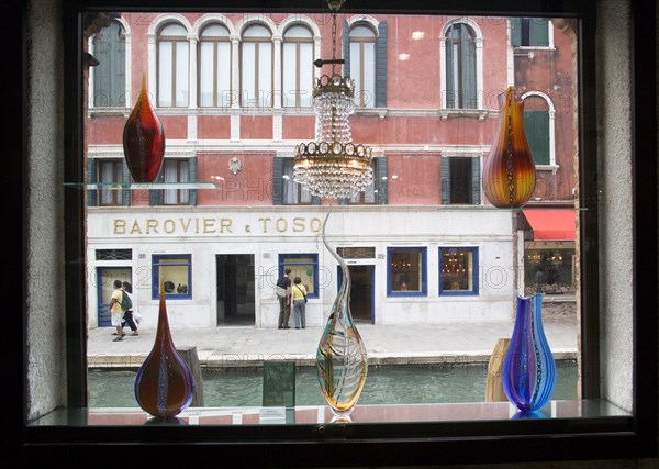 ITALY, Veneto, Venice, Tourists walking along the Fondamenta dei Vetrai looking in the windows of a glass shop gallery beside the Rio dei Vetrai canal on the lagoon island of Murano. Seen through a window display of modern blown glass