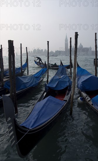 ITALY, Veneto, Venice, A gondolier works his gondola past others moored at the Molo San Marco with Palladio's church of San Giorgio Maggiore on the island of the same name in the distance on a misty day