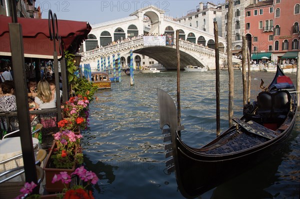 ITALY, Veneto, Venice, A gondola moored beside a restaurant on the Grand Canal with tourists dining. Beyond the Rialto Bridge lined with tourists spans the canal