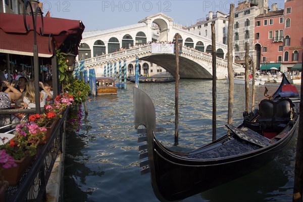 ITALY, Veneto, Venice, A gondola moored beside a restaurant on the Grand Canal with tourists dining. Beyond the Rialto Bridge lined with tourists spans the canal