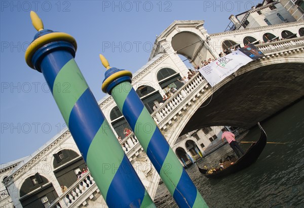 ITALY, Veneto, Venice, Colourful posts for mooring boats in front of gondolas carrying sightseers on the Grand Canal beneath the Rialto Bridge lined with tourists